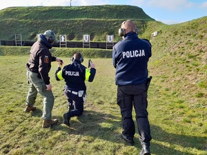 na wojskowej strzelnicy instruktor dokonuje przeglądu broni i magazynka, które trzyma w uniesionych rękach policjant. Obok niego stoi drugi instruktor