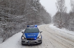 oznakowany policyjny radiowóz na ośnieżonej otoczonej lasem drodze