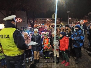 wieczorowa pora. W centrum miasta policjant ruch drogowego, trzymając w dłoni opaski odblaskowe rozmawia z dziećmi