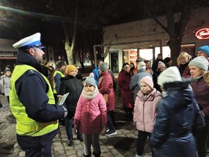 wieczorowa pora. W centrum miasta policjant ruch drogowego, trzymając w dłoni opaski odblaskowe rozmawia z dziećmi