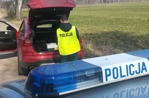 policjant stoi przy otwartym bagażniku samochodu osobowego. Za nim zaparkowany jest oznakowany radiowóz