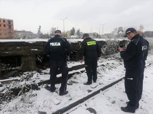 przy starej ośnieżonej wyładowni PKP stoją dwaj umundurowani policjanci i strażnik miejski. Pod rampą siedzą bezdomne osoby