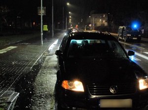 po zmierzchu. Tuż za przejściem dla pieszych stoi vw passat. Za nim znajduje się policjant w kamizelce odblaskowej. Po przeciwnej stronie, wzdłuż jezdni stoi nieoznakowany pojazd z włączonym światłem błyskowym