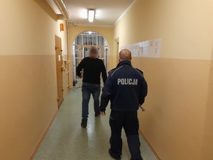 korytarzem pomieszczenia dla osób zatrzymanych idzie mężczyzna, za nim idzie policjant służby dyżurnej trzymając w ręce klucze do celi