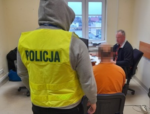 w pokoju przesłuchań obok biurka przy którym znajduje się nieumundurowany policjant siedzi na krześle zatrzymany mężczyzna z rękoma skutymi kajdankami. Za nim stoi policjant w kamizelce odblaskowej z napisem policja