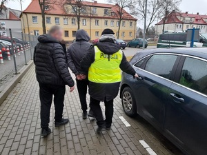 dwaj policjanci prowadzą w stronę nieoznakowanego radiowozu mężczyznę skutego kajdankami