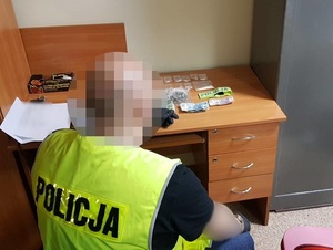 nieumundurowany policjant w kamizelce z napisem policja siedzi przed biurkiem, na którym znajdują się worek z suszem roślinnym, woreczki oraz zawiniątka z proszkiem i kryształkami oraz banknoty