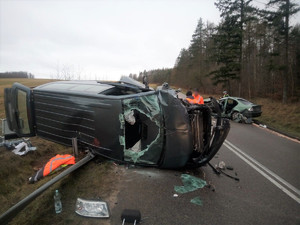 miejsce wypadku drogowego. Częściowo na jezdni i na poboczu leży przewrócony na bok vw transporter z rozbitą szybą i licznymi uszkodzeniami karoserii. Po drugiej stronie drogi znajduje się uszkodzone osobowe bmw, przy którym stoi policjant ruchu drogowego