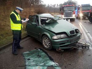 miejsce wypadku drogowego. Przy zielonym osobowym bmw z licznymi uszkodzeniami stoi policjant ruchu drogowego, który wykonuje oględziny. Za nim na drodze stoją dwa wozy straży pożarnej