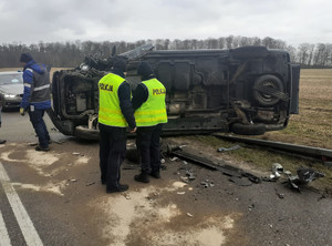 miejsce wypadku drogowego. Przy leżącym na boku na drodze samochodzie typu bus stoją dwaj policjanci ruchu drogowego