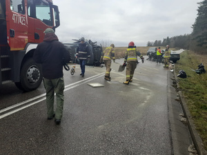 miejsce wypadku drogowego. Po obu stronach drogi znajdują się uszkodzone samochody. Jeden, typu bus leży na boku. Technik kryminalistyki mierzy odległość, strażacy niosą pojemnik, przy zielonym bmw stoi policjantka, która sporządza oględziny