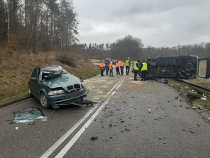 miejsce wypadku drogowego. Po obu stronach drogi znajdują się uszkodzone pojazdy: zielone osobowe bmw i leżący na boku pojazdy typu bus. Przy busie stoją dwaj policjanci ruchu drogowego, obok przechodzi kolejny. Na drodze stoją mężczyźni w pomarańczowych odblaskowych kurtkach. Przed bmw leży na drodze rozbita szyba