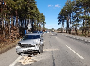 miejsce kolizji drogowej. Na poboczu stoją jeden za drugim samochody osobowe. Pierwszy ma uszkodzony przód. Za nimi znajduje się policyjny radiowóz