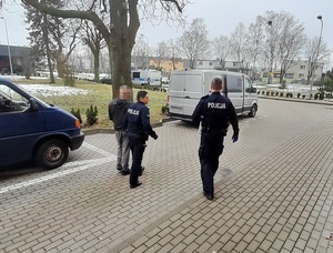 policjant służby konwojowej prowadzi mężczyznę do nieoznakowanego radiowozu. Obok nich idzie drugi policjant