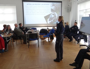 w sali klubu osiedlowego policjantka przemawia przez mikrofon do seniorów siedzących przy stole. Obok wyświetla się przygotowana przez policjantów prezentacja na temat oszustw.