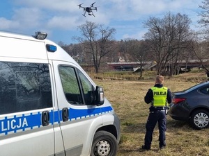 na polu obok radiowozu stoi policjant który spogląda w górę na dron