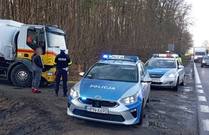 na drodze podporządkowanej, prostopadle do jezdni stoi samochód ciężarowy. Przy nim stoi kierowca oraz policjant ruchu drogowego. Na poboczu wzdłuż jezdni stoją dwa oznakowane policyjne radiowozy