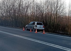 częściowo na jezdni i na poboczu znajduje się srebrny vw lupo z uszkodzonym tyłem. Jest ogrodzony słupkami