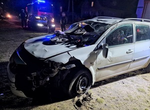 Pora nocna. Uszkodzone białe renault z licznymi wgnieceniami na karoserii, pękniętymi szybami, zerwanym lusterkiem i częścią zderzaka. W tle wóz straży pożarnej i policjant ruchu drogowego