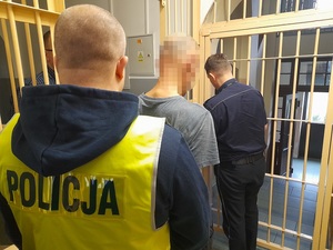 policjant w kamizelce odblaskowej z napisem policja stoi za zatrzymanym mężczyzną. Okratowane drzwi pomieszczenia dla osób zatrzymanych otwiera policjant służby dyżurnej