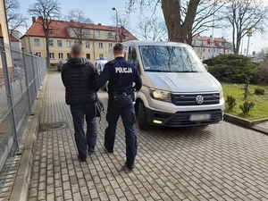 na wewnętrznym placu komendy policji policjant prowadzi skutego kajdankami mężczyznę w kierunku nieoznakowanego radiowozu