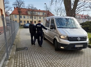 na wewnętrznym placu komendy policji policjant prowadzi skutego kajdankami mężczyznę do nieoznakowanego radiowozu