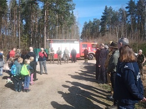 na drodze leśnej stoją zgromadzone osoby, wśród nich m.in. leśnicy, duchowny, policjantka