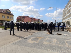 policjanci w strojach ochronnych do zabezpieczania imprez masowych otaczają grupę pseudokibiców zgromadzonych na osiedlu. Wśród policjantów znajdują się psy służbowe