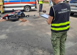 miejsce wypadku drogowego. Na ulicy leży uszkodzony motocykl, kilka metrów za nim leży kask motocyklisty. Na miejscu zdarzenia jest karetka pogotowia, policjant ruchu drogowego i technik kryminalistyki, który przeprowadza oględziny