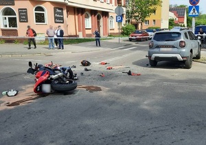 miejsce wypadku drogowego odgrodzone taśmą policyjną. Na skrzyżowaniu ulic leży uszkodzony motocykl. Przed nim, częściowo na chodniku stoi dacia duster. Przy motocyklu widać ślady wycieków płynów eksploatacyjnych. Na jezdni leżą fragmenty motocykla