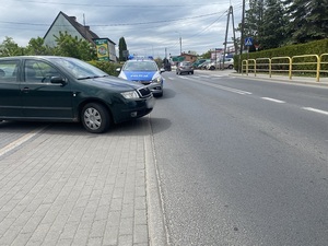 na chodniku stoi ciemnozielona skoda fabia. Za nią znajduje się oznakowany policyjny radiowóz