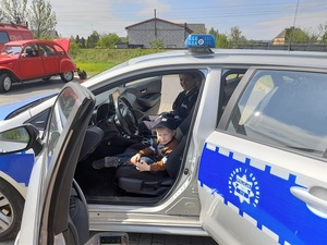 w policyjnym radiowozie na miejscu kierowcy siedzi mały chłopiec z policyjną czapką na głowie. Obok niego siedzi policjantka