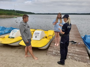 nad jeziorem policjantka wręcza ulotki młodym dziewczynie i chłopakowi stojącym przy rowerach wodnych