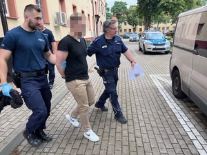 dwaj policjanci prowadzą zatrzymanego mężczyznę w kierunku nieoznakowanego radiowozu