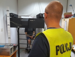 w pomieszczeniu techników kryminalistyki przed aparaturą do robienia zdjęć siedzi zatrzymany mężczyzna. Przed nim stoi policjant