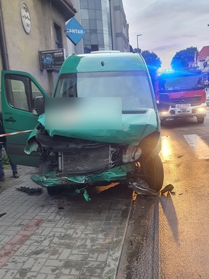 Na chodniku i częściowo na jezdni przy przejściu dla pieszych znajduje się zielony mercedes sprinter z uszkodzonym przodem. Za nim stoi wóz straży pożarnej