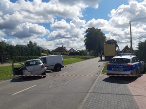 miejsce wypadku drogowego odgrodzone taśmą. Na jezdni i poboczu znajdują się dwa samochody, które stykają się ze sobą. Toyota ma otwarte drzwi od strony kierowcy. Na jezdni leżą odłamki części samochodów. Dalej po przeciwnej stronie jezdni stoi samochód ciężarowy, przy którym stoją dwie osoby: policjant i mężczyzna. Przed ciężarówką znajduje się policyjny radiowóz