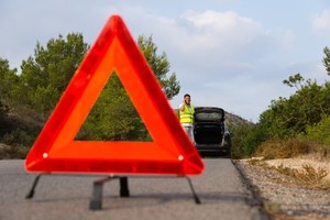 na pierwszym planie znajduje się trójkąt ostrzegawczy ustawiony na jezdni. W tle widać samochód z otwartą klapą bagażnika oraz mężczyznę w kamizelce odblaskowej