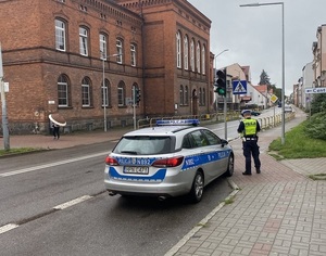 obok szkoły przed przejściem dla pieszych z sygnalizacją świetlną przy oznakowanym radiowozie stoi policjant ruchu drogowego.