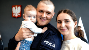 w auli uśmiechnięty umundurowany policjant w stopniu posterunkowego trzyma na rękach niemowlę. Obok niego stoi uśmiechnięta młoda kobieta. W tle widać godło Polski