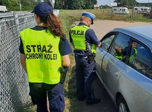 policjant ruchu drogowego rozmawia z kierowcą osobowego samochodu. Za policjantem stoi funkcjonariuszka Straży Ochrony Kolei