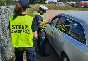 policjant ruchu drogowego kieruję w stronę kierowcy siedzącego w samochodzie osobowym apteczkę pierwszej pomocy. Za policjantem stoi funkcjonariuszka Straży Ochrony Kolei