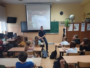 klasa szkolna. Policjant rozmawia z uczniami. Za nim wyświetla się prezentacja multimedialna