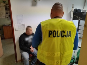 w pomieszczeniu technika kryminalistyki siedzi na krześle zatrzymany mężczyzna z rękoma skutymi kajdankami. Przed nim stoi policjant