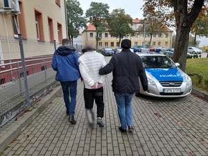 nieumundurowany policjant prowadzi mężczyznę skutego kajdankami w kierunku radiowozu