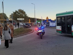 policjant jadący motocyklem z włączonymi sygnałami błyskowymi. Przez jezdnię przechodzą piesi, którzy kierują się w stronę cmentarza