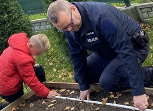 policjant mierzy miarką odległość właściwą do umieszczania w donicy sadzonek. Obok niego znajduje się chłopiec, który przytrzymuje miarkę