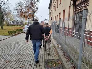 dwaj nieumundurowani policjanci prowadzą mężczyznę skutego kajdankami w kierunku wejścia do budynku komendy