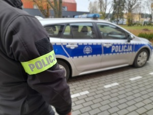 na tle oznakowanego radiowozu stoi policjant kryminalny. Na rękawie kurtki ma opaskę z napisem Policja