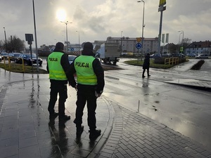 Przed przejściem dla pieszych stoją dwaj policjanci ruchu drogowego. Przez pasy przechodzi kobieta